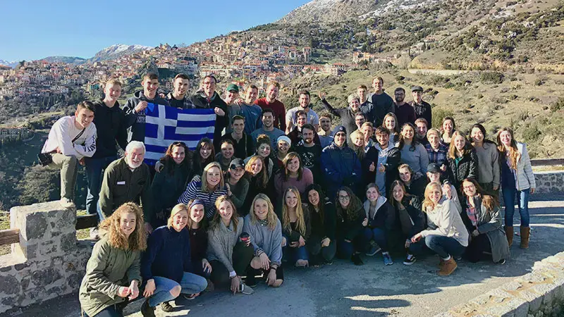 A group of students and faculty standing by a historic city while holding the Greek flag during a J-term trip