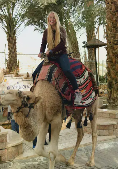 A blonde female student in front of palm trees riding a camel