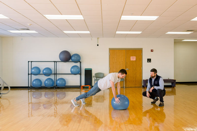A professor watching a student do a push up on an exercise ball