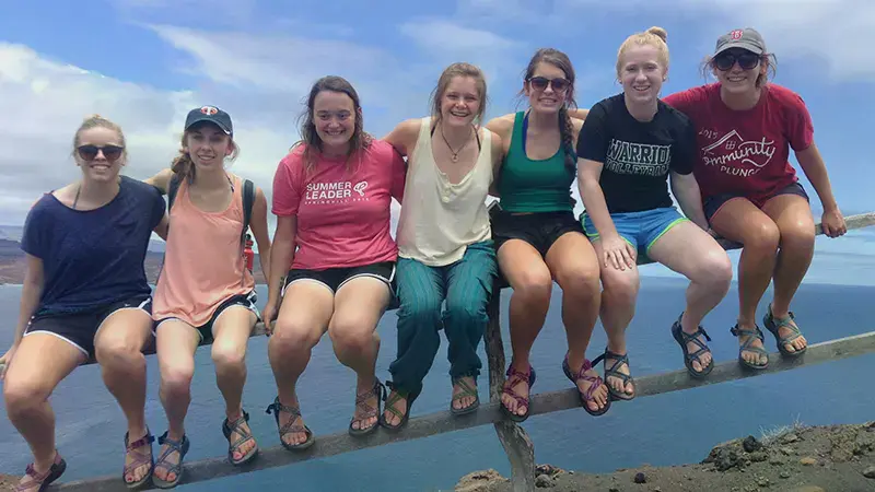 A group of female students sitting on an iron railing that separates them and steep fall into the ocean